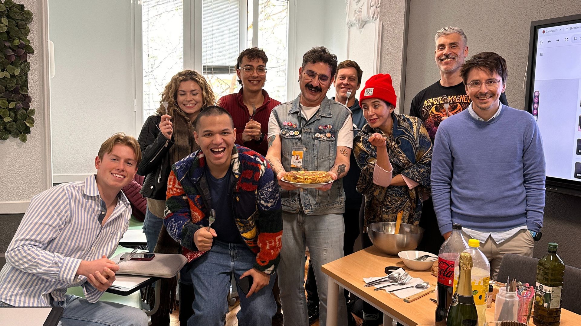 A diverse group of happy students and a teacher in a classroom after making a traditional Spanish tortilla, smiling and posing for the camera with a plate of food and cooking supplies.