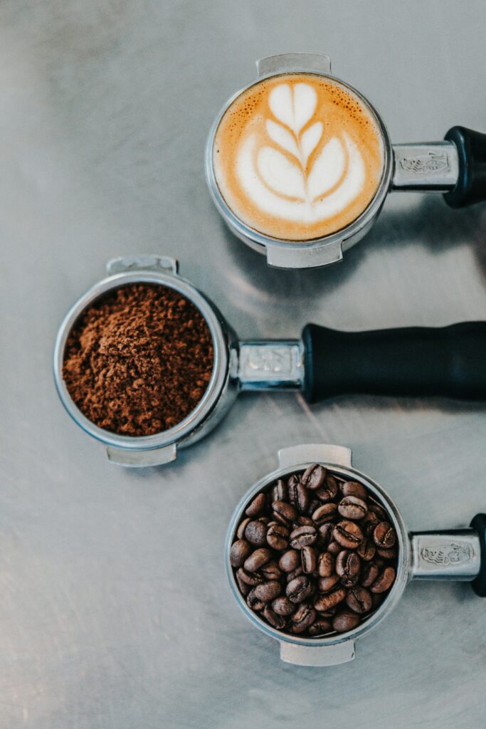 Three metal portafilters showing coffee beans, ground coffee, and a finished cappuccino with latte art — representing Spanish coffee culture and café traditions in Madrid.