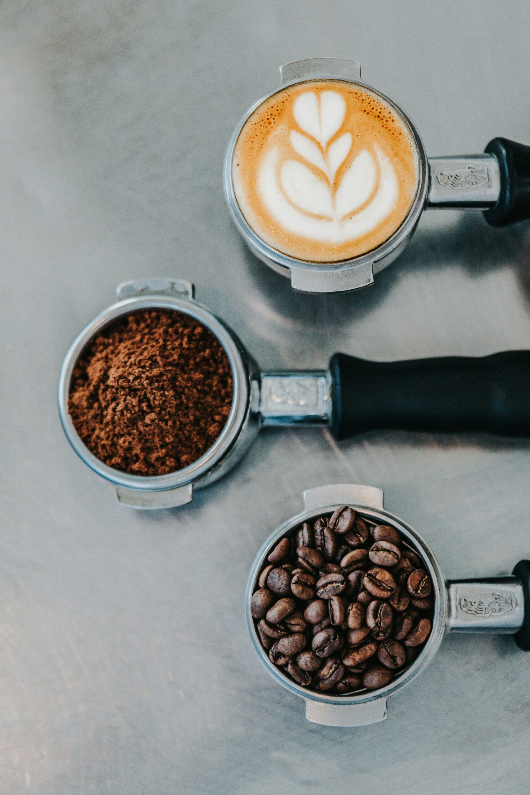 Three metal portafilters showing coffee beans, ground coffee, and a finished cappuccino with latte art — representing Spanish coffee culture and café traditions in Madrid.