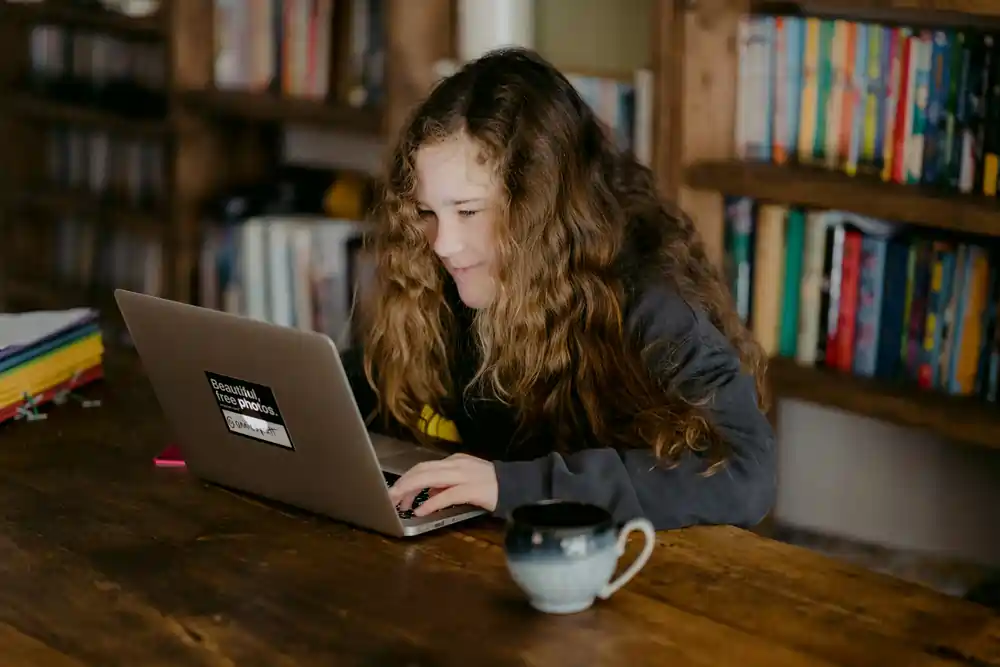 A young person with long curly hair using a laptop at a wooden table in front of bookshelves, practicing Spanish immersion through digital devices.