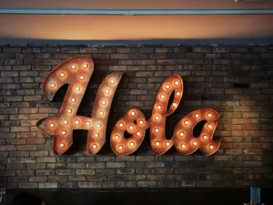 A vintage-style neon "Hola" sign on a brick wall in a Madrid cafe, representing the first step in learning Spanish and building confidence.