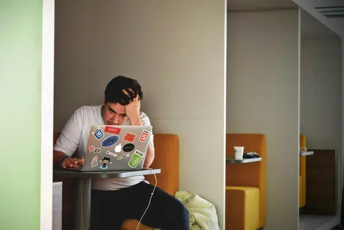 A man looking stressed and holding his head while working on a laptop in an outdoor Madrid setting, representing the frustration of navigating life in Spain with only English.