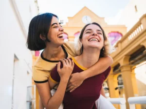 Two young women laughing joyfully outdoors in Madrid, one giving the other a piggyback ride, representing friendship and a sense of belonging.