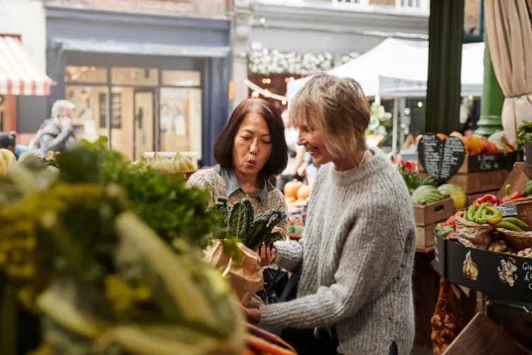 Two mature women, one with dark hair and one with blonde hair, smiling and discussing produce while shopping at a vibrant Madrid street market.