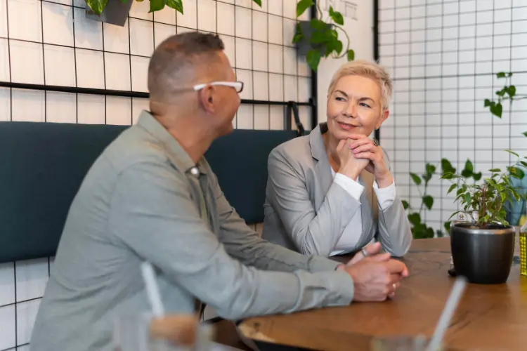 A mature man and woman sitting at a wooden table in a stylish cafe with plants, engaged in a warm and focused conversation over coffee.