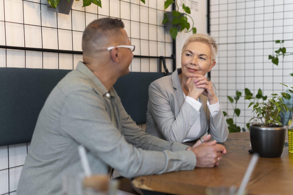 Un hombre y una mujer de edad madura sentados a una mesa de madera en una cafetería elegante, entablando una conversación cálida y enfocada mientras toman café.