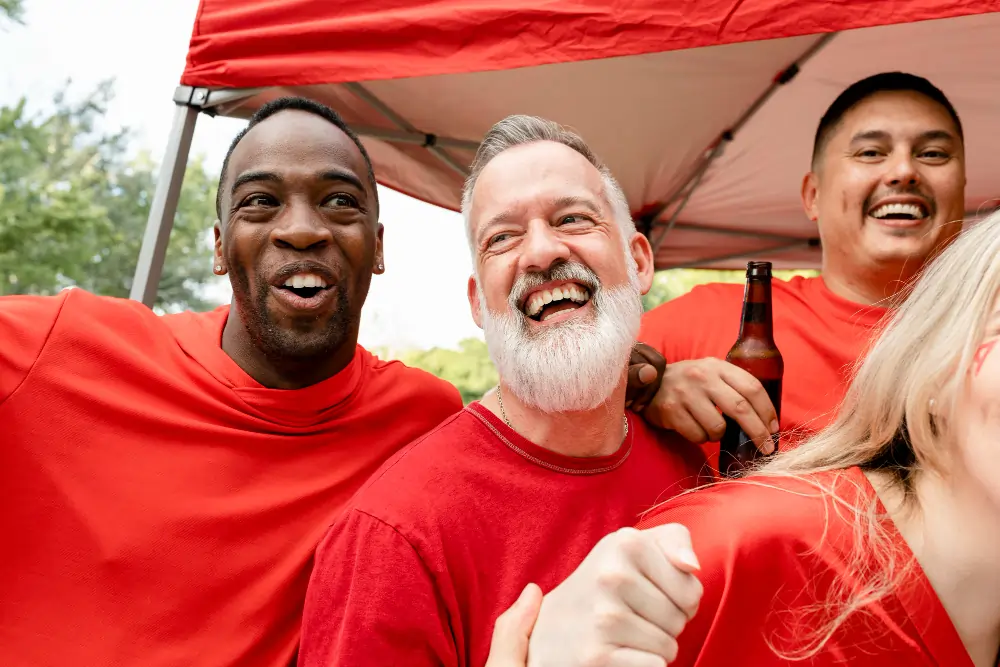 A diverse group of cheerful friends wearing bright red t-shirts laughing, cheering, and holding a beer bottle together under an outdoor red canopy.
