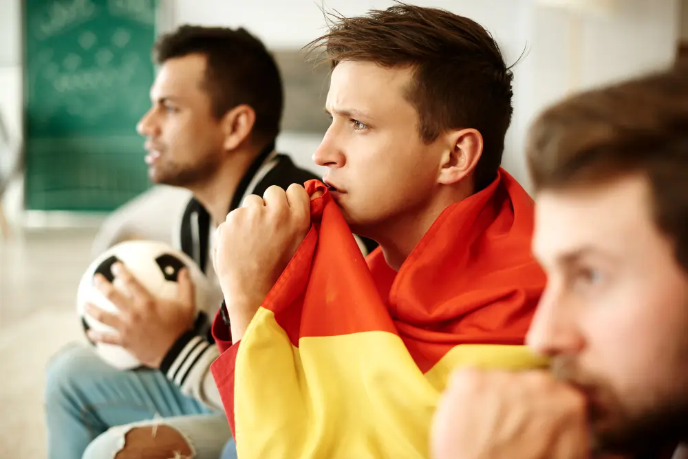 A close-up side view of three young men watching a football match intensely, with the central man holding a red and yellow Spanish flag to his mouth in suspense.
