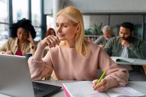 A woman taking handwritten notes at her laptop in a focused study session with other adult students in the background