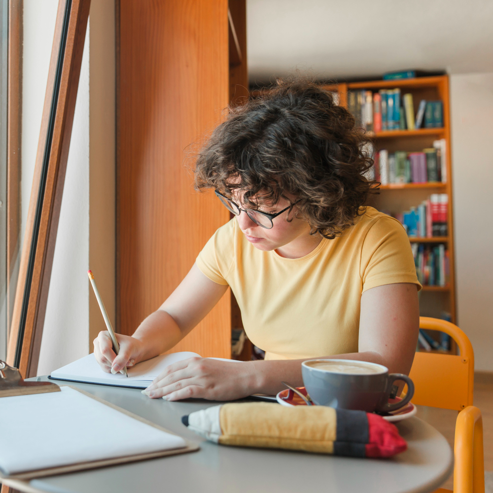 A student sitting at a desk with a coffee cup, focused on writing and studying with pencil and paper near a bookshelf.