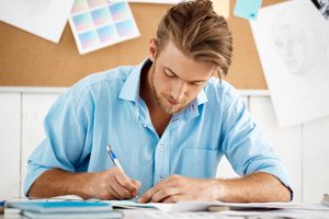 A young man in a blue shirt concentrating as he takes notes and plans his study timeline at a desk.