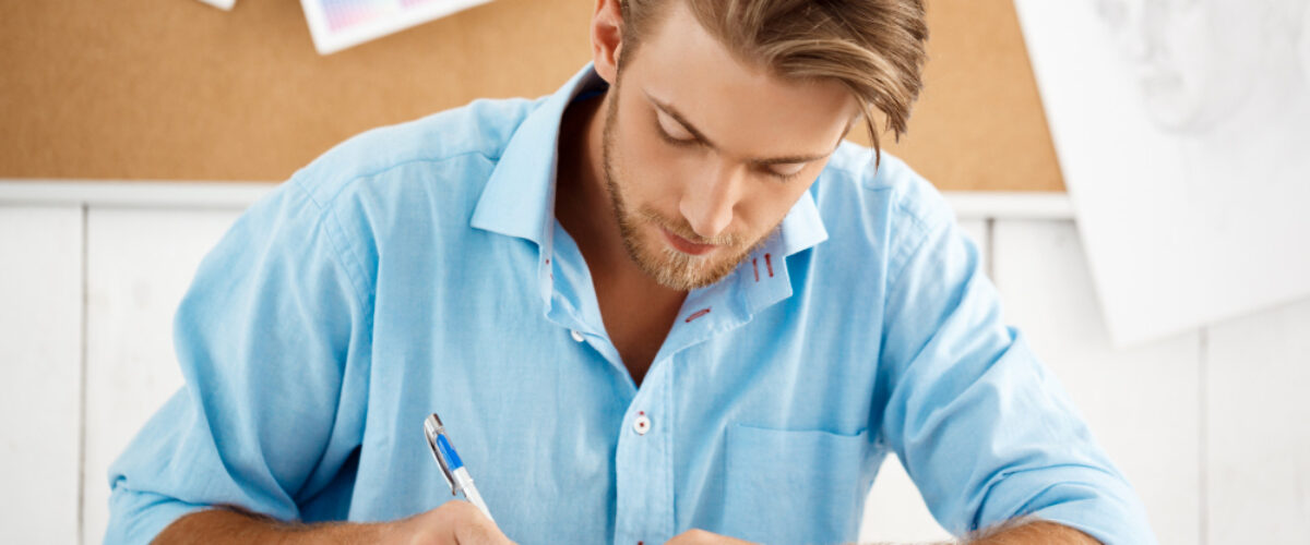 Un hombre joven con camisa azul concentrado mientras toma notas y planifica su calendario de estudio en un escritorio.