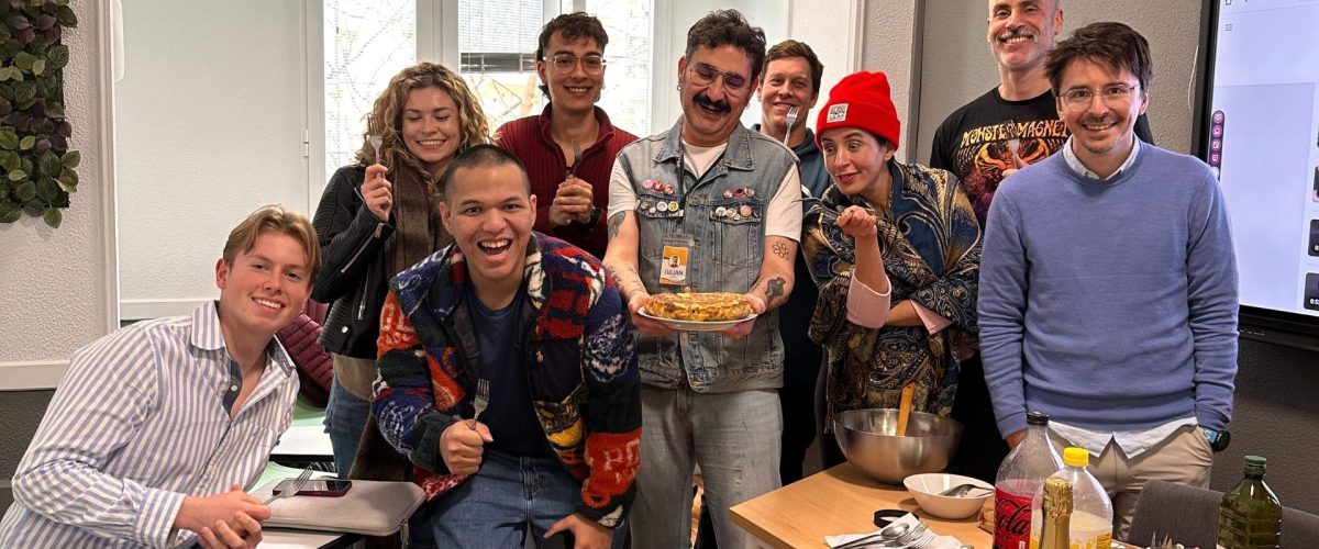 A diverse group of happy students and a teacher in a classroom after making a traditional Spanish tortilla, smiling and posing for the camera with a plate of food and cooking supplies.