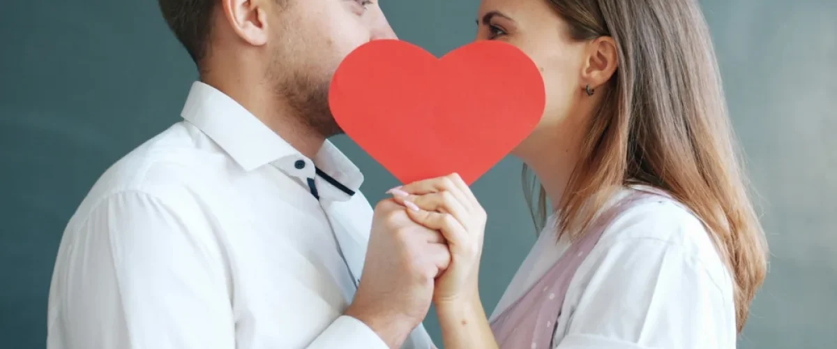 Un hombre y una mujer con camisas blancas sostienen un corazón de papel rojo entre ellos, simbolizando la naturaleza emocional y romántica del idioma español.