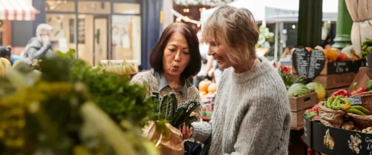 Dos mujeres adultas de pie en un mercado luminoso, mirando de cerca un manojo de verduras de hoja verde y participando en una conversación expresiva.