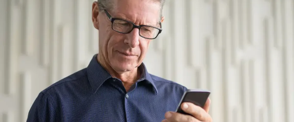 A mature man with glasses in a blue shirt looking intently at his smartphone screen, practicing Spanish vocabulary through an app.
