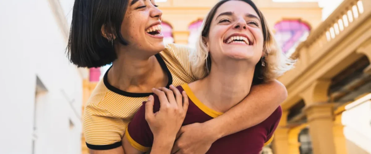 Dos mujeres jóvenes riendo alegremente al aire libre en Madrid, una cargando a la otra a caballito, representando la amistad y el sentido de pertenencia.