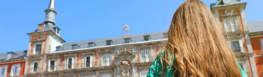 Young woman with long hair standing in front of Plaza Mayor in Madrid, looking up at the historic architecture on a sunny day